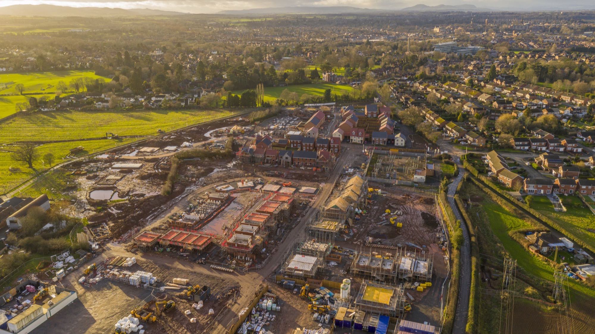 Aerial view at sunset of a new housing development under construction on the edge of an English town, with open farmland on one side and an established estate on the other.